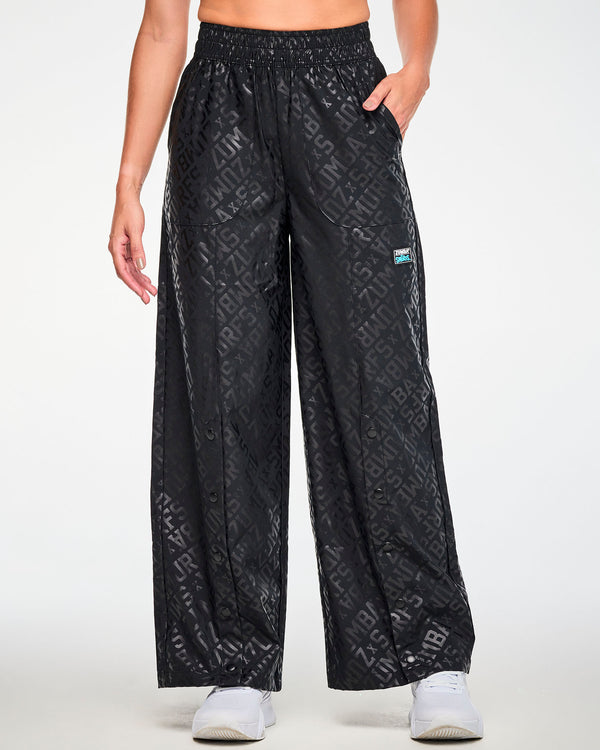 Female model with one hand in the pocket of the black tonal logo print wide-leg pants, showing the elastic waistband, black snaps down the front of each leg, and a black patch with white 'ZUMBA' and blue 'THE SMURFS' logos on the left hip. Paired with white shoes.