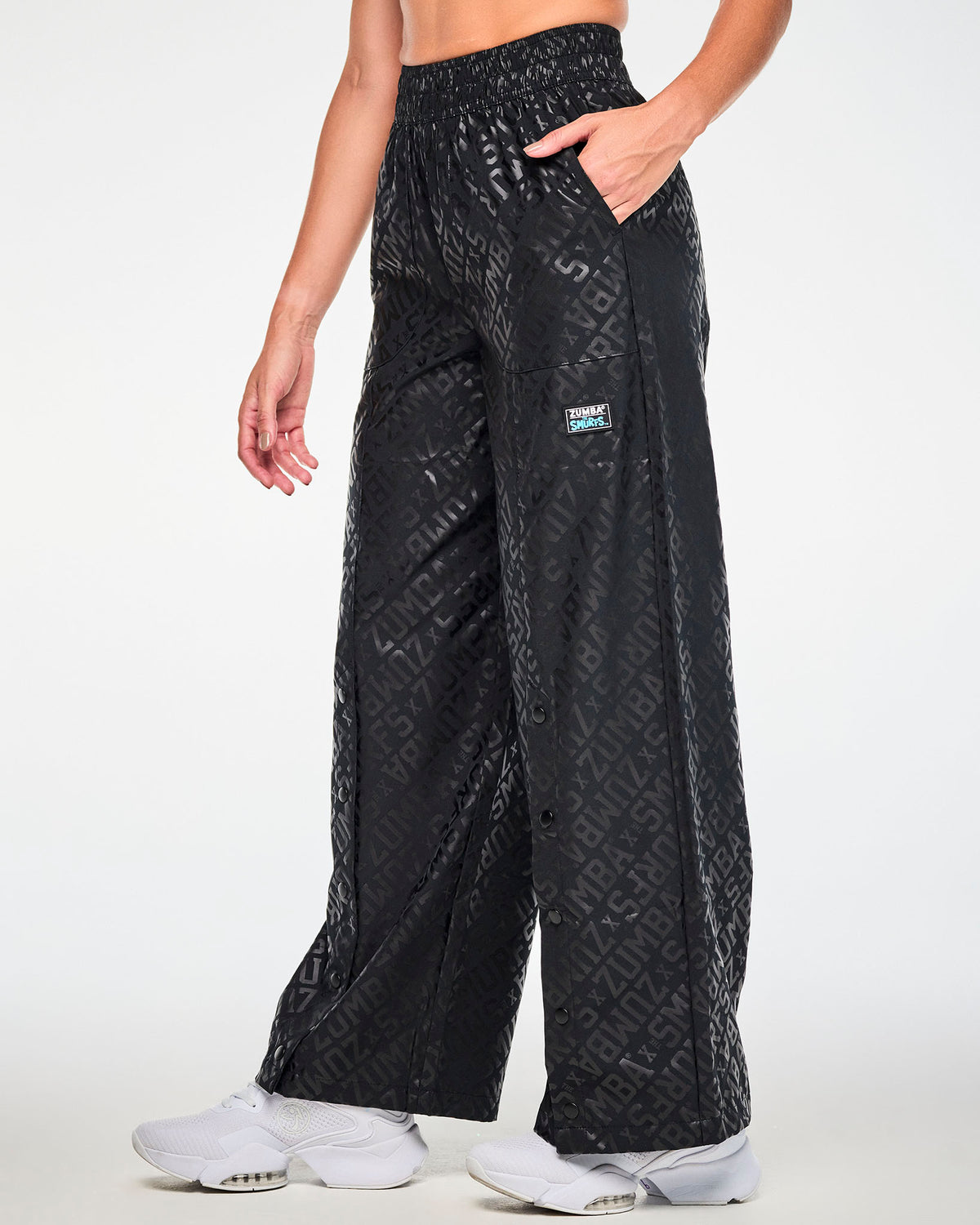 Female model in black wide-leg pants with a glossy tonal all-over print of diagonal 'ZUMBA' X 'THE SMURFS' text, shown in side view with the row of black snaps running down the leg and the logo patch on the left hip.