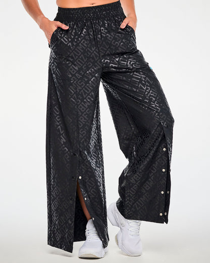 Female model with both hands in the pockets of the black tonal logo print wide-leg pants, with the snaps undone near the hem of each leg to open a front slit.