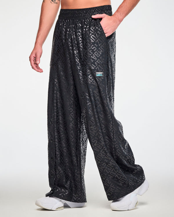 Male model in black wide-leg pants with a glossy tonal print of diagonal 'ZUMBA' X 'THE SMURFS' text, shown in side view mid-step with the snaps running down each leg and the black 'ZUMBA' X 'THE SMURFS' patch on the left hip.
