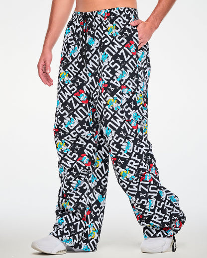Male model with one hand in the pocket of oversized cargo pants with an all-over print of bold white diagonal 'ZUMBA' X 'THE SMURFS' text on black, scattered with Papa Smurf in red, Smurfette with yellow hair, and other blue Smurfs, shown in side view with the flap cargo pocket and a black drawcord at the hem.