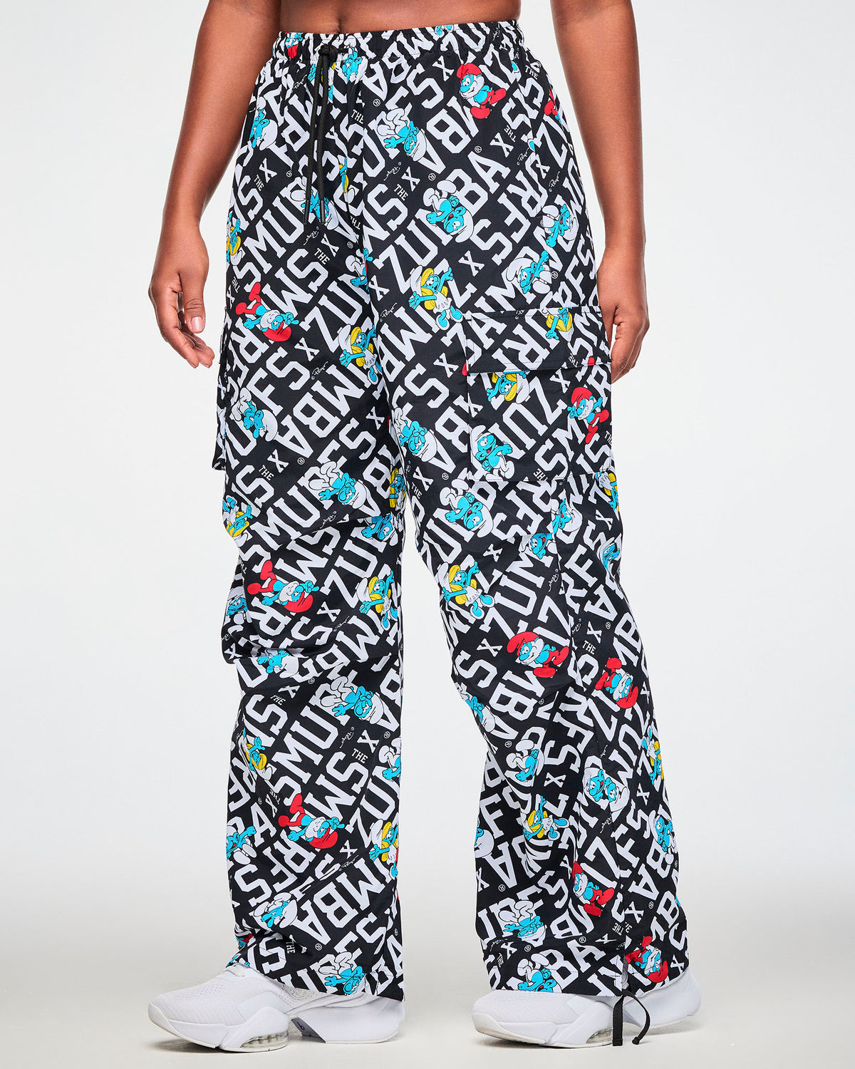Female model in oversized cargo pants printed all over with bold white diagonal 'ZUMBA' X 'THE SMURFS' text on black, with Papa Smurf, Smurfette, and other blue Smurfs, shown in side view with the relaxed leg and a flap cargo pocket on the outer thigh.