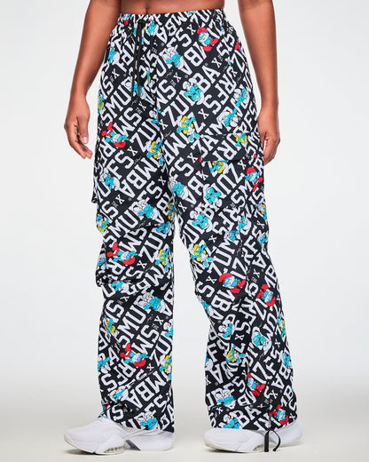 Female model in oversized cargo pants printed all over with bold white diagonal 'ZUMBA' X 'THE SMURFS' text on black, with Papa Smurf, Smurfette, and other blue Smurfs, shown in side view with the relaxed leg and a flap cargo pocket on the outer thigh.