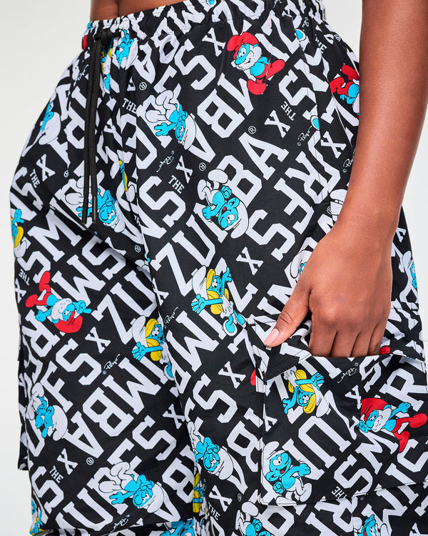 Close-up of the black and white Smurfs print cargo pants, showing Papa Smurf, Smurfette, and a smiling Smurf among white block letters spelling 'ZUMBA' X 'THE SMURFS', with small Peyo signatures, the black waist drawcord, and a flap cargo pocket.
