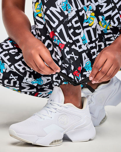 Close-up of a female model tying the black drawcord at the hem of the black and white Smurfs print cargo pants to cinch the ankle, above a white shoe.