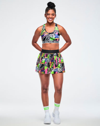 Full view of a female model wearing a skort with a vibrant graffiti-style print in neon green, pink, yellow, and black, paired with green and white socks and white sneakers.
