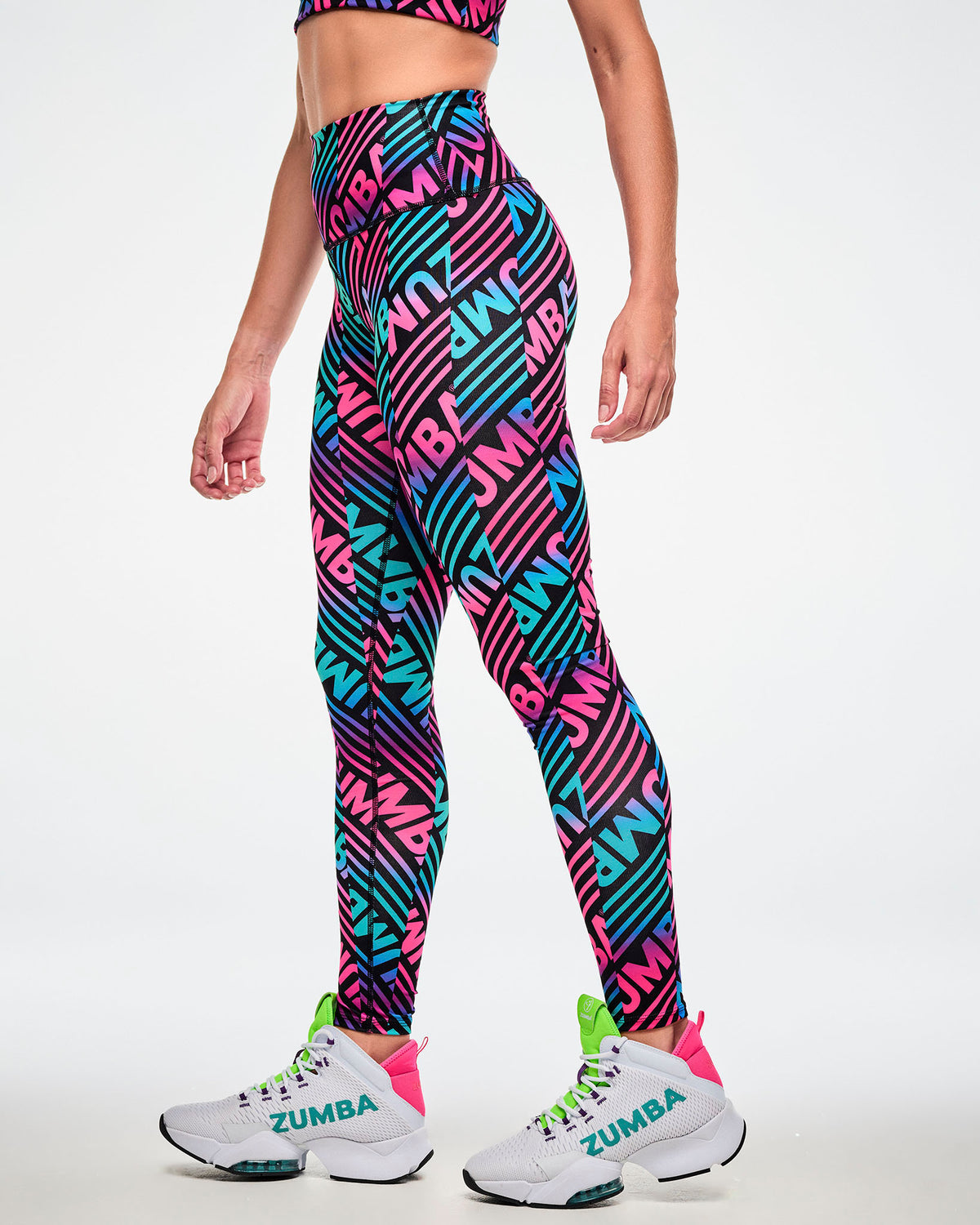 Female model wearing high-waisted ankle leggings with a repeated pink, aqua, and black "ZUMBA" print, paired with white and green shoes, side view.