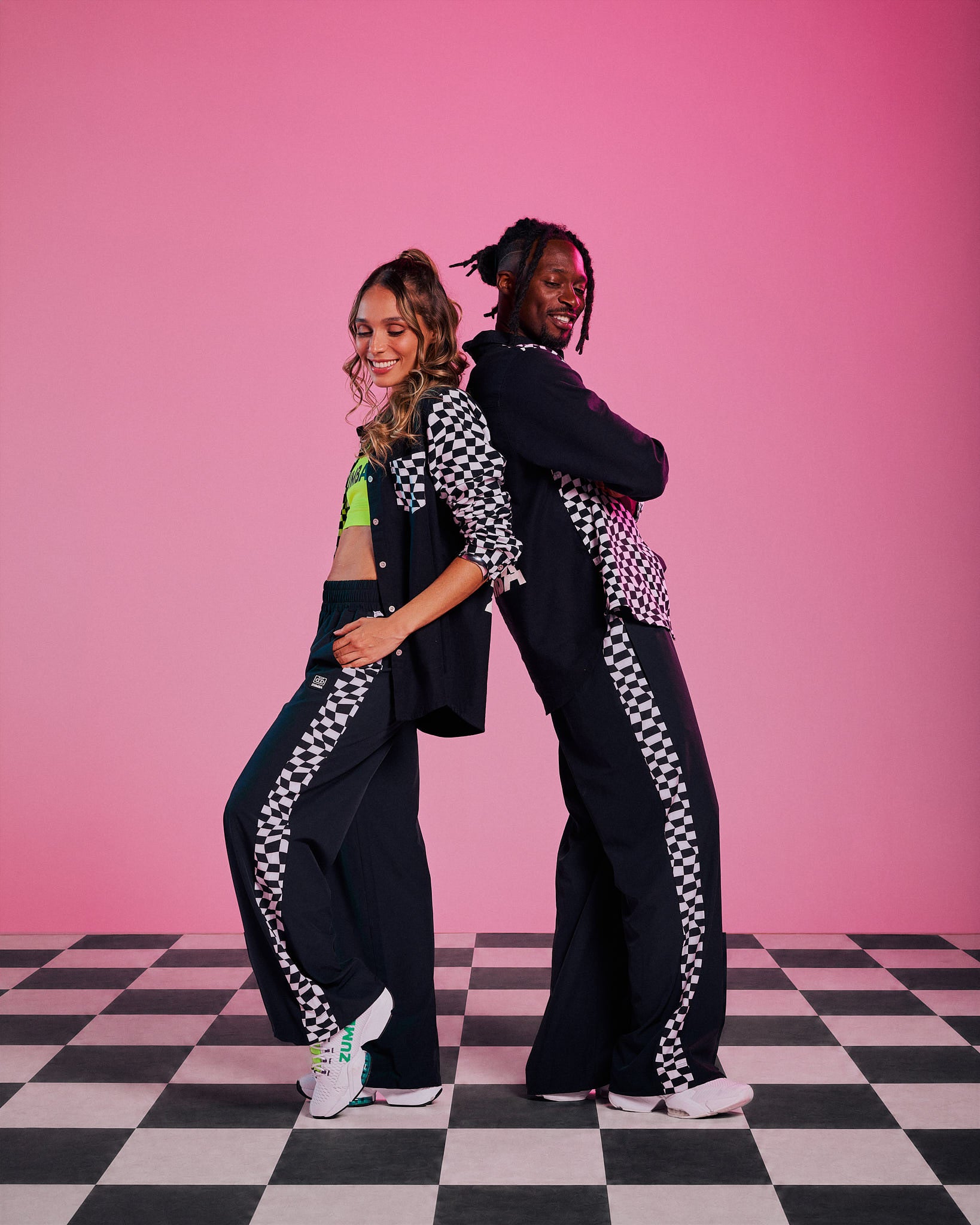 Two models (female and male), posing back-to-back in matching black track pants with white and black checkered side stripes, paired with black checkered long-sleeve tops, against a solid pink background and a black-and-white checkered floor.