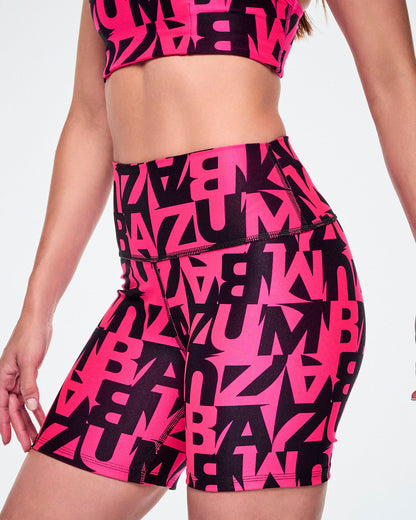 A close-up of a female model wearing high-waisted biker shorts and a matching sports bra with a bold black and bright pink abstract ZUMBA letter print.