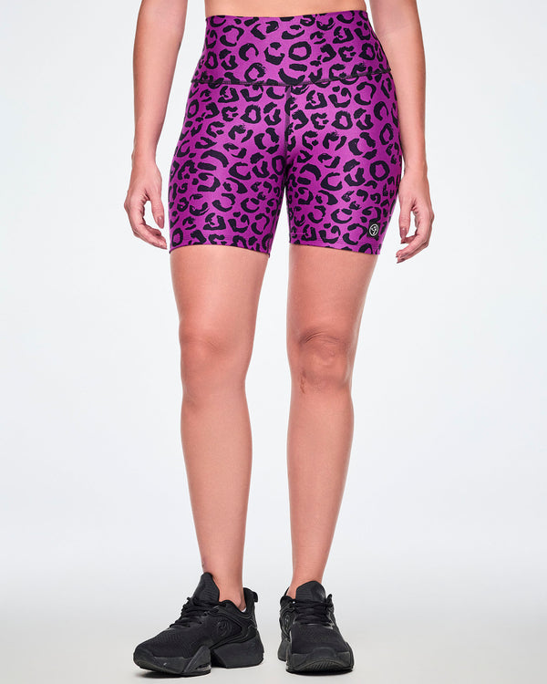 Front view of a female model wearing high-waisted purple biker shorts with a black leopard print pattern and a small Zumbito logo near the hem and paired with black shoes.