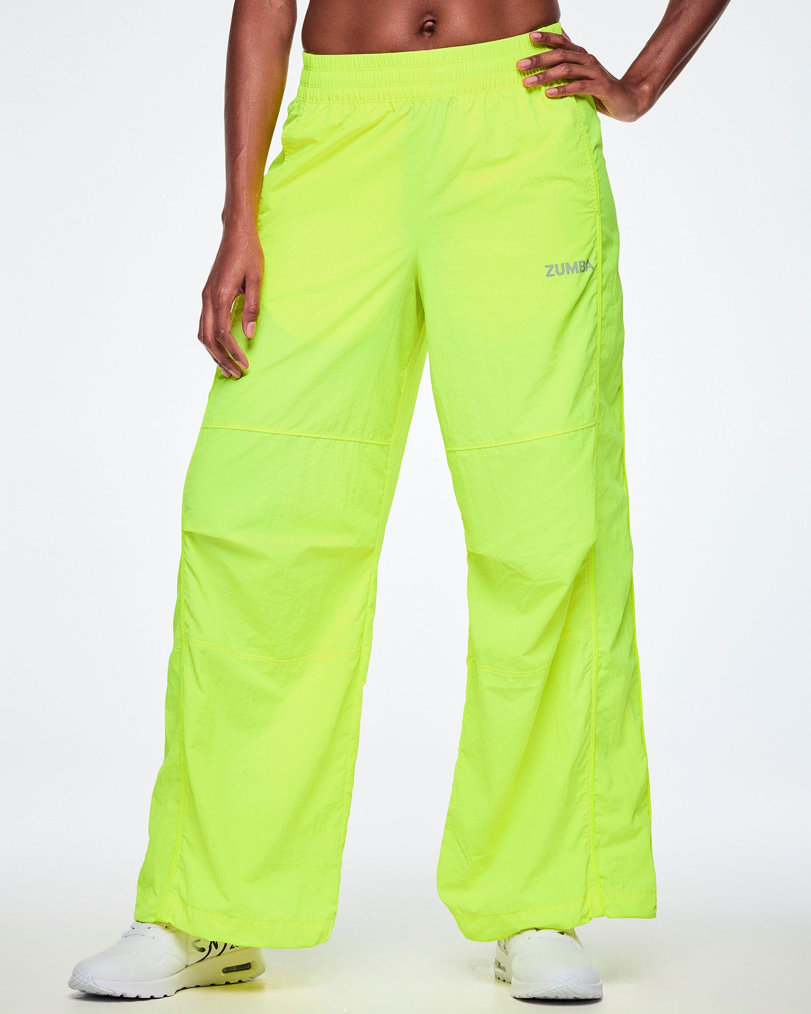 Front view of a female model wearing bright neon yellow-green wide-leg cargo pants with a small silver ZUMBA text on the left thigh, paired with white shoes.