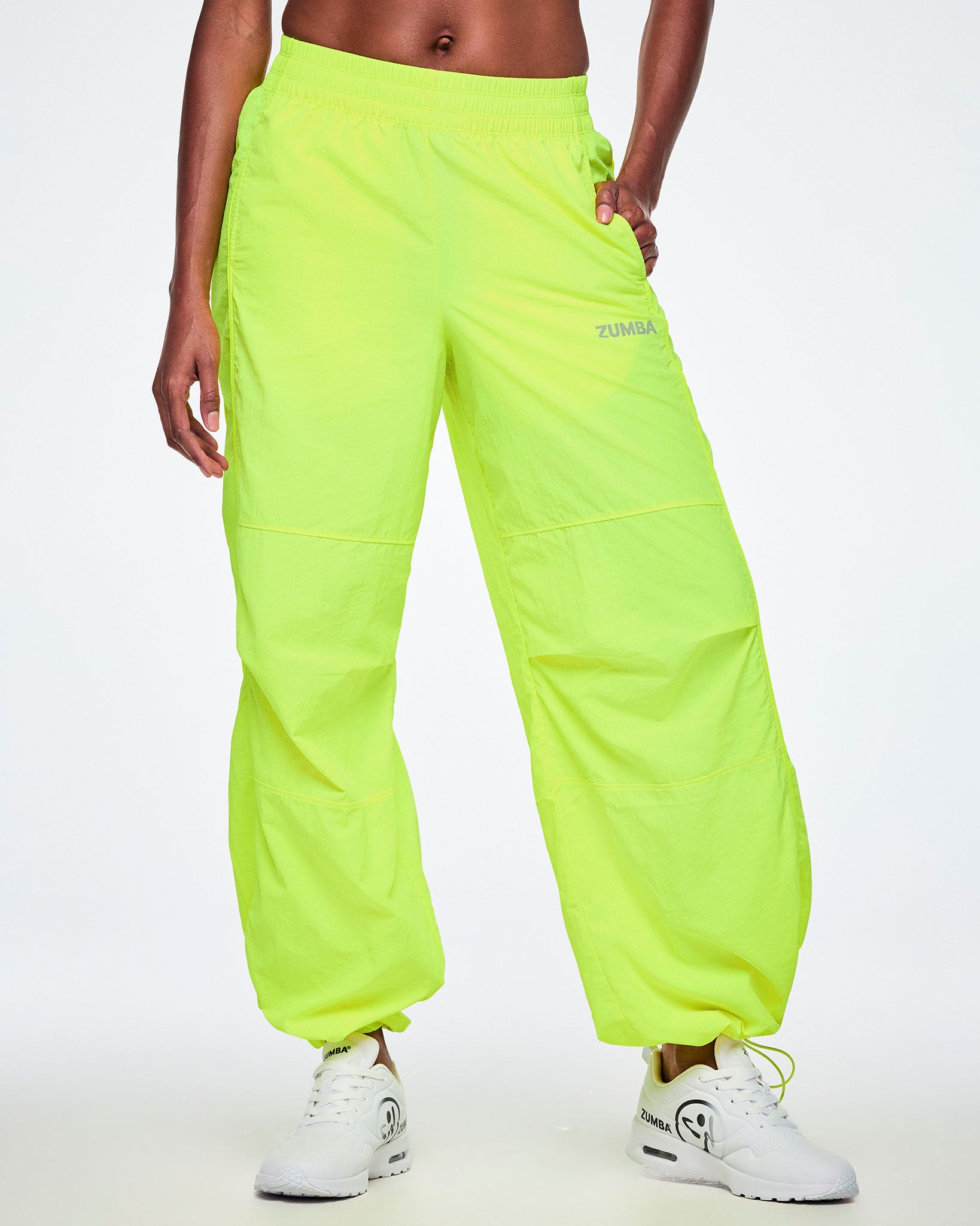 Front view of a female model wearing bright neon yellow-green wide-leg cargo pants with her hands in her pockets, showing more of the small silver ZUMBA text on the left thigh and paired with white shoes.