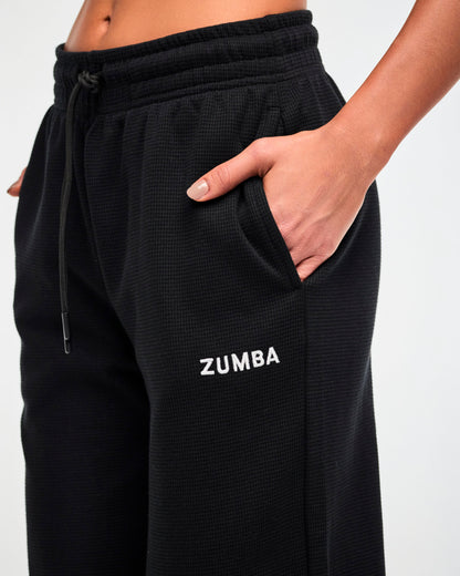 Close-up of the drawstring waistband and white 'ZUMBA' text on the black waffle-knit track pants, with a hand tucked into the side pocket.