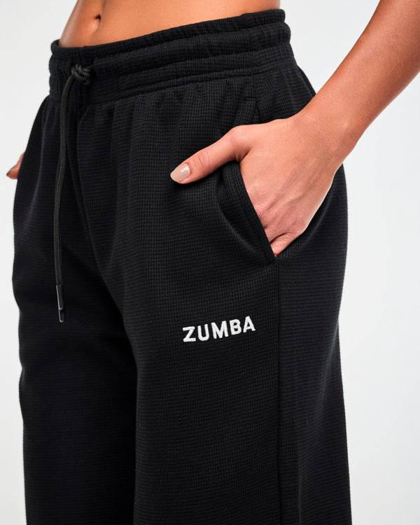 Close-up of the drawstring waistband and white 'ZUMBA' text on the black waffle-knit track pants, with a hand tucked into the side pocket.