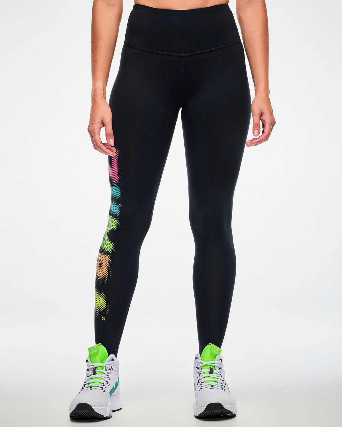 Female model, front view, wearing black high-waisted ankle leggings with a partial view of the dot-gradient "ZUMBA" text down the right leg and white shoes with a green tongue and purple accents.