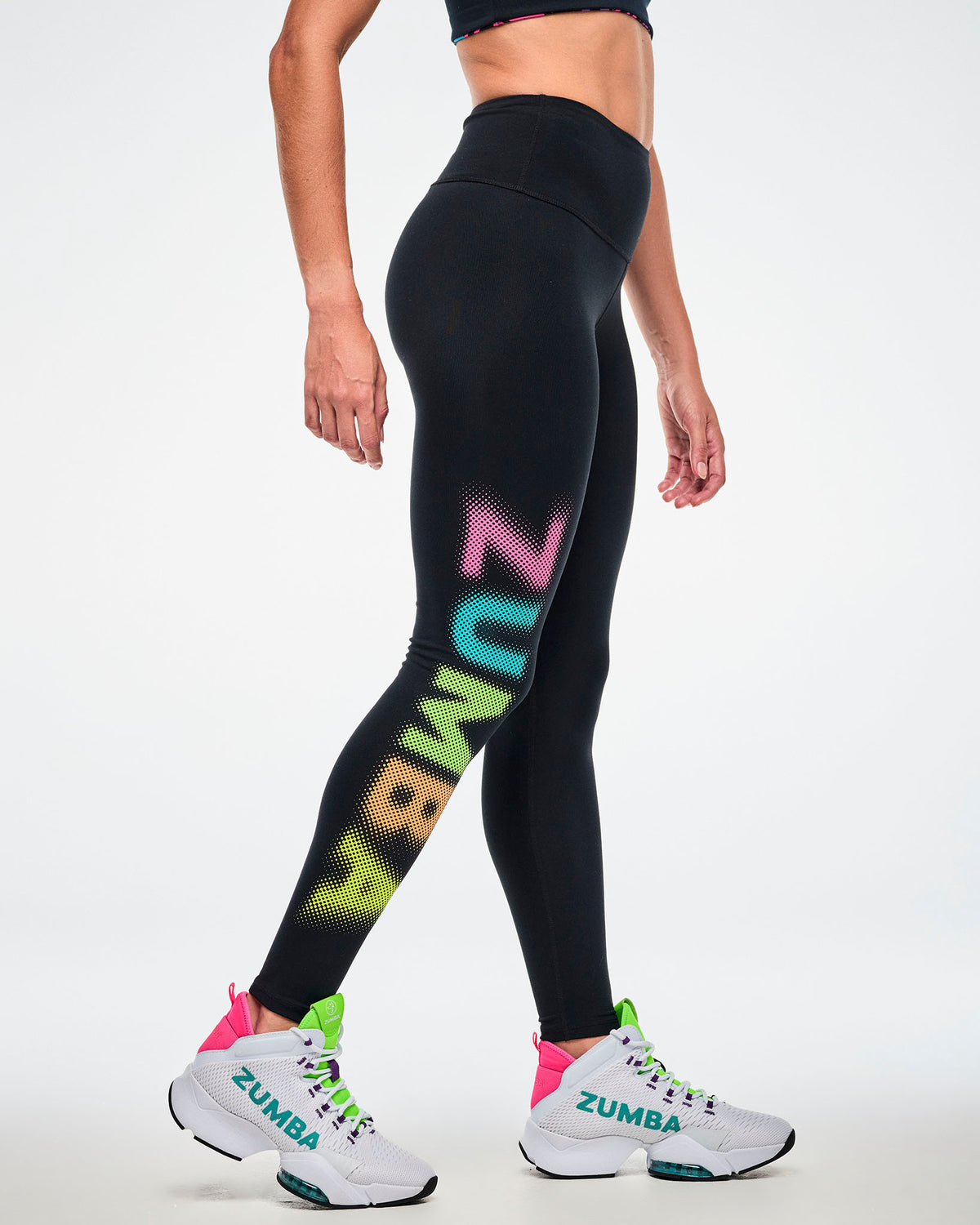 Female model wearing black high-waisted ankle leggings with a dot-gradient graphic "ZUMBA" text, in pink, aqua, green, orange and yellow text down the right leg, paired with white shoes.