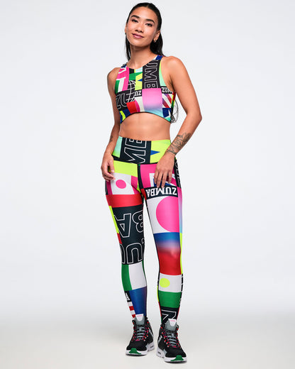 Female model wearing a colorful sports bra and matching leggings, paired with black Zumba shoes against a white background.