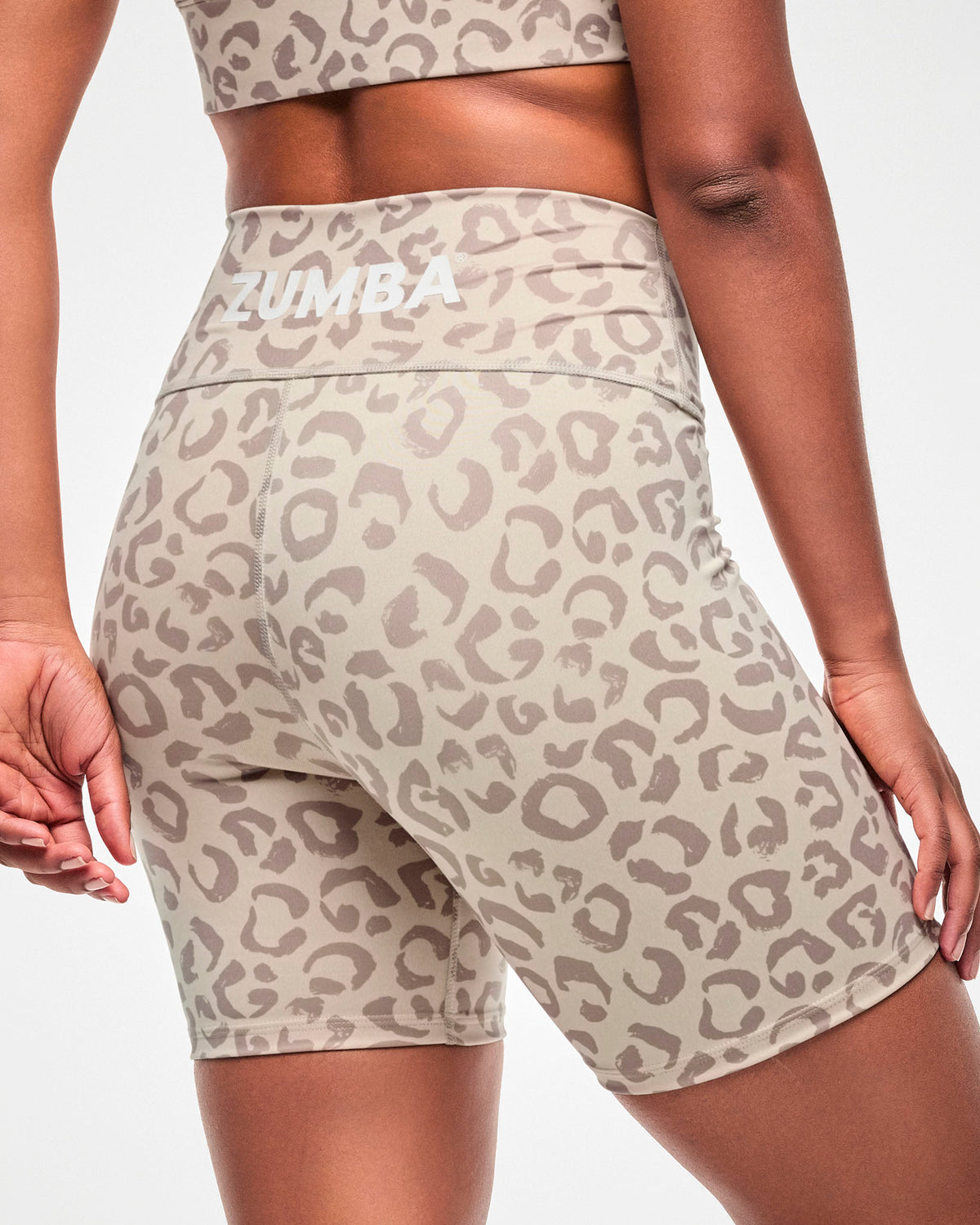 Close-up back view of beige high-waisted biker shorts with a light brown leopard print and white 'ZUMBA' text across the back waistband.
