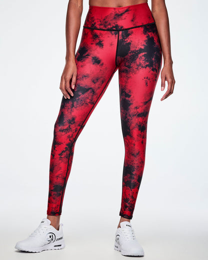 Front view of a female model wearing high-waisted red and black tie-dye like leggings, paired with white shoes with a big black Zumbito faded logo and black Zumba text.