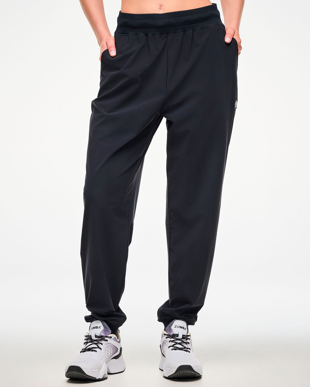 Front view of a female model wearing high waisted black jogger pants with hands in the side pockets and paired with white shoes.