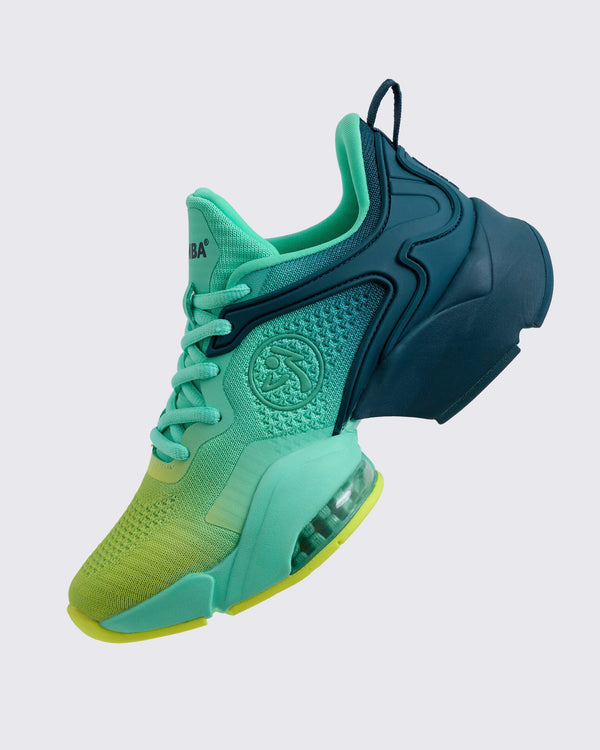 Side view of the gradient teal-to-yellow Zumba sneaker showing the embossed circular logo on the mid foot, mint laces, navy heel cage, and clear air unit in the midsole, on a light gray background.