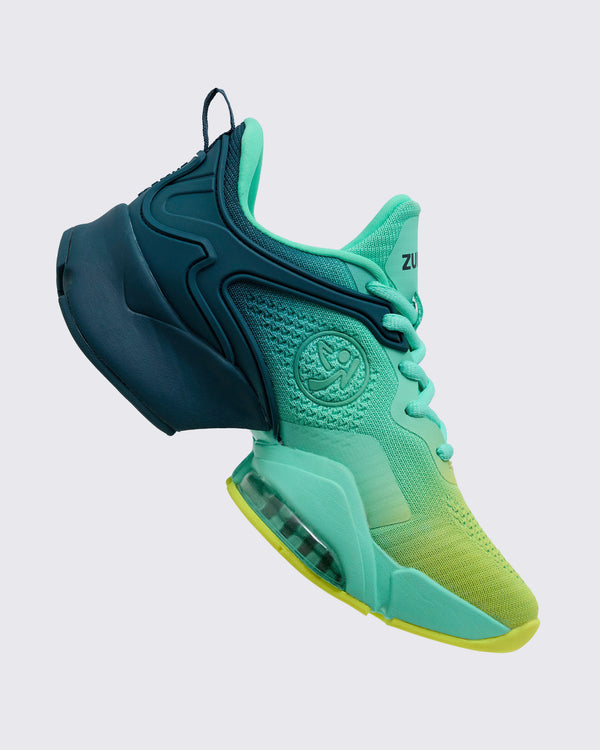 Side view of the gradient teal-to-yellow Zumba sneaker showing the embossed circular logo on the mid foot, mint laces, navy heel cage, and clear air unit in the midsole, on a light gray background.