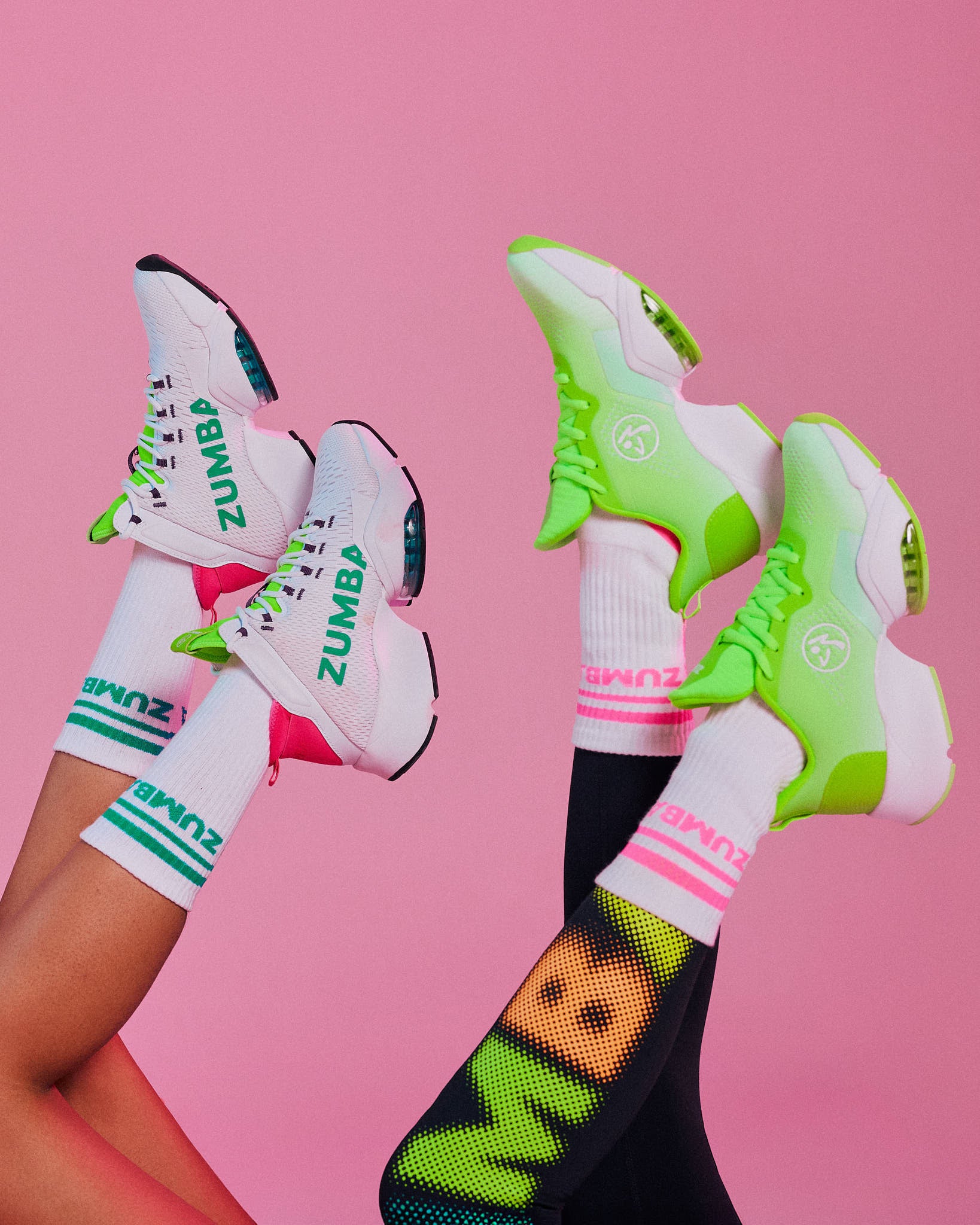 Two pairs of feet raised wearing a white "ZUMBA" sneaker with a black sole and a neon green sneaker with a white sole, against a pink background.