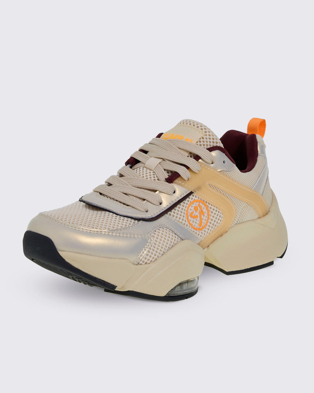 Front side view of a beige platform shoe with a mesh upper, metallic gold and translucent tan overlays, and burgundy lining. Details include an orange Zumbito logo, orange 'ZUMBA' text on the tongue, an orange heel pull tab, and a chunky split sole with a clear air cushion.