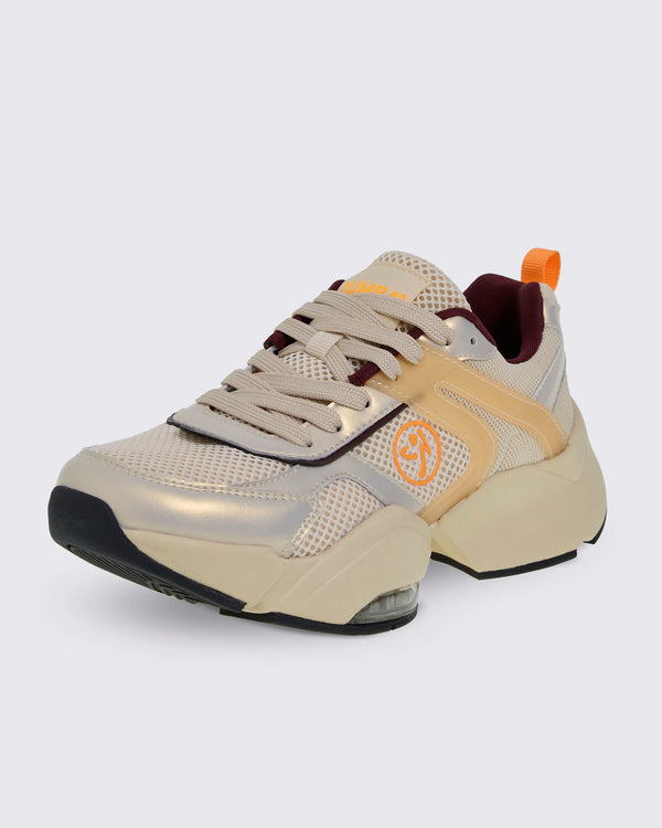 Front side view of a beige platform shoe with a mesh upper, metallic gold and translucent tan overlays, and burgundy lining. Details include an orange Zumbito logo, orange 'ZUMBA' text on the tongue, an orange heel pull tab, and a chunky split sole with a clear air cushion.