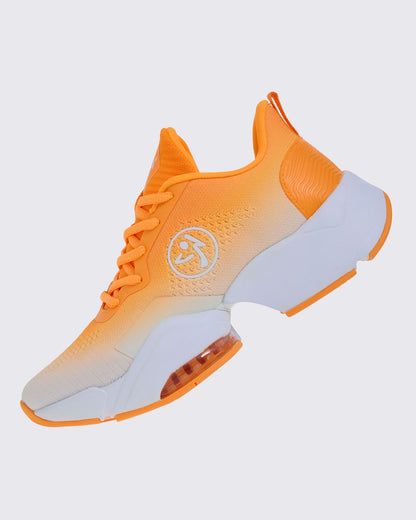 Left side view of the shoe, highlighting the orange-to-white ombre knit, a white Zumbito logo, a wavy textured orange heel panel, and a clear air cushion under the forefoot.