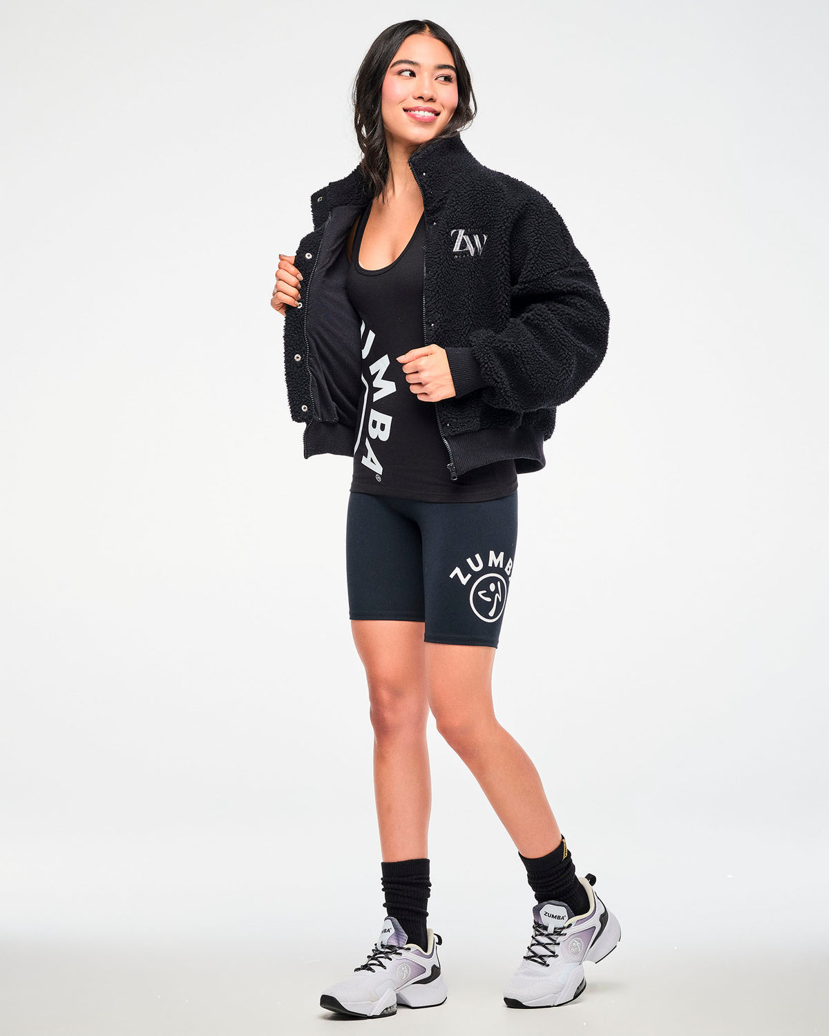 Full-body view of a female model wearing a black sherpa jacket with "ZW" embroidered logo, layered over a "ZUMBA" graphic tank top, paired with black Zumba biker shorts, socks, and white shoes.