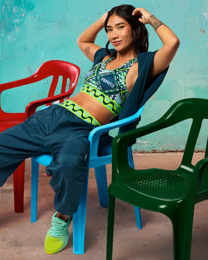 Green printed Zumba sports bra with wide lime wave band and racerback fit. Female model lounges in teal jogger pants with a teal jacket draped over her shoulders and teal-to-yellow ombre sneakers, posed on colorful plastic chairs against a teal wall.