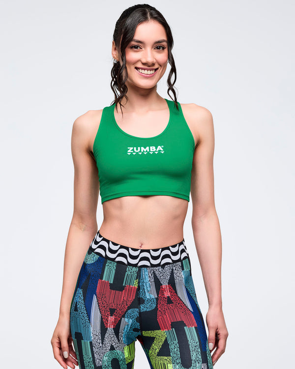 Front view of a green Zumba scoop neck tank with white logo, styled with multicolor graphic leggings featuring a black-and-white wavy waistband; female model stands on a white background.