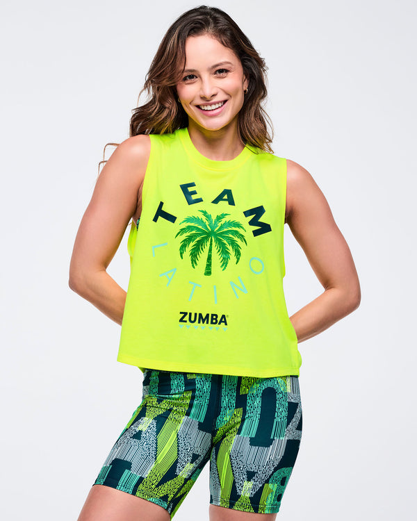 Front view of a neon yellow “TEAM LATINO” sleeveless Zumba tank with palm tree graphic, styled with green-and-black abstract print biker shorts on a smiling female model against a white background.