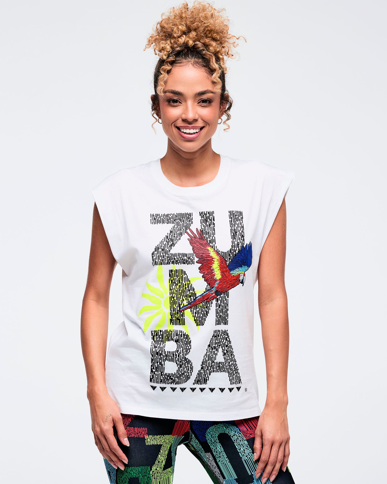 White sleeveless muscle tee with large “ZUMBA” text graphic and a colorful parrot and palm print on the front, worn by a female model with multicolor patterned leggings.
