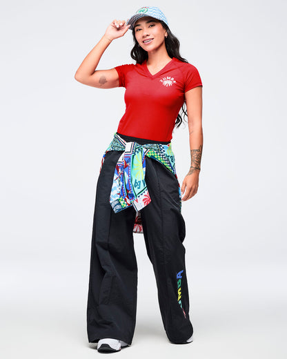 Red fitted V-neck Zumba tee with small white palm logo on the left chest, styled with loose black wide-leg pants, a colorful graphic button-down, tied at the waist, a blue-and-white patterned baseball cap, and white sneakers; female model poses on a white background.