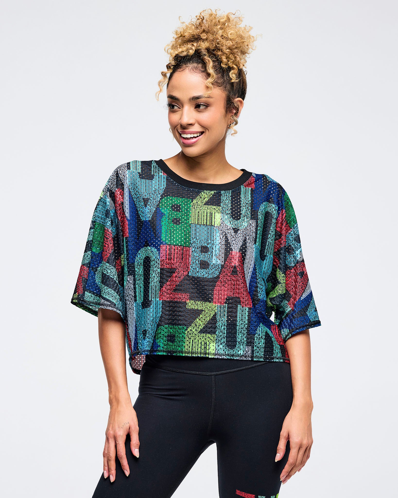 Multicolor Zumba mesh crop top with oversized fit, short sleeves, and black ribbed crew neck, covered in abstract “ZUMBA” lettering. Female model pairs it with high-waisted black leggings on a light gray studio background.