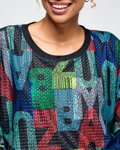 Close-up of the multicolor Zumba mesh crop top with bold “ZUMBA” lettering print and black ribbed crew neckline. Female model is shown in a close-up wearing the textured, breathable top on a light gray studio background.