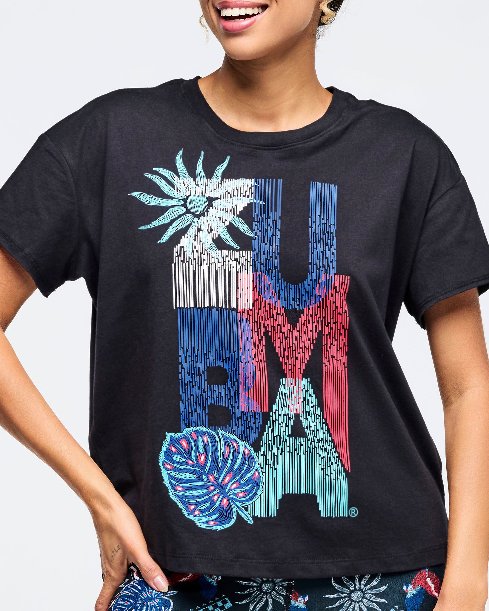 Close-up of a black short-sleeve crewneck tee with oversized “ZUMBA” vertical graphic in blue, red, teal and white with tropical flower and leaf accents; female model shown from chest to hips wearing matching dark tropical-print leggings on a white background.