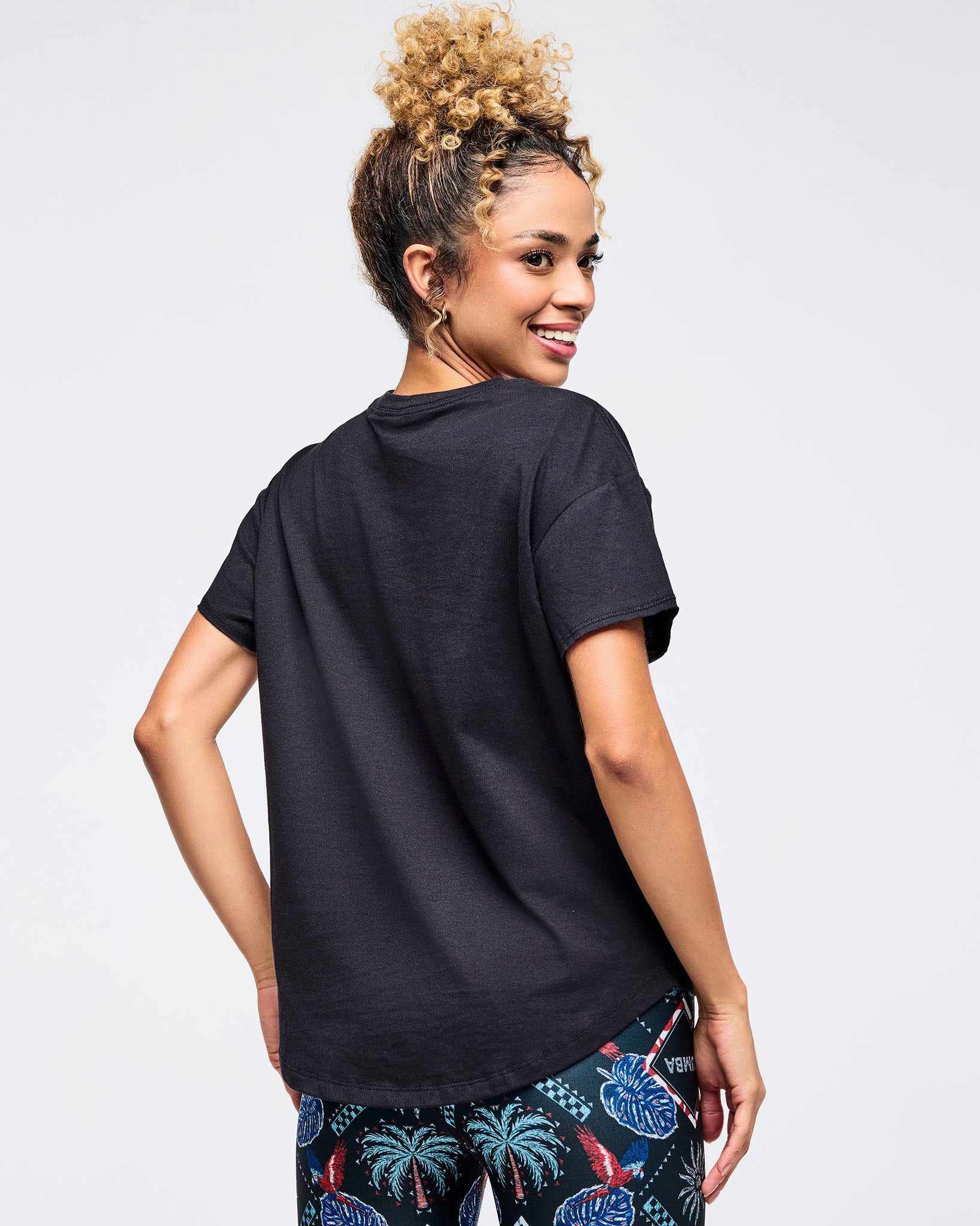 Back view of a black relaxed-fit crewneck tee on a female model, paired with dark tropical-print leggings featuring palm trees and floral motifs, photographed on a white background.