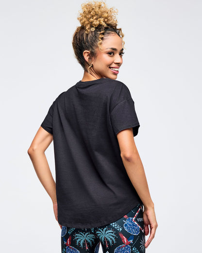 Back view of a black relaxed-fit crewneck tee on a female model, paired with dark tropical-print leggings featuring palm trees and floral motifs, photographed on a white background.