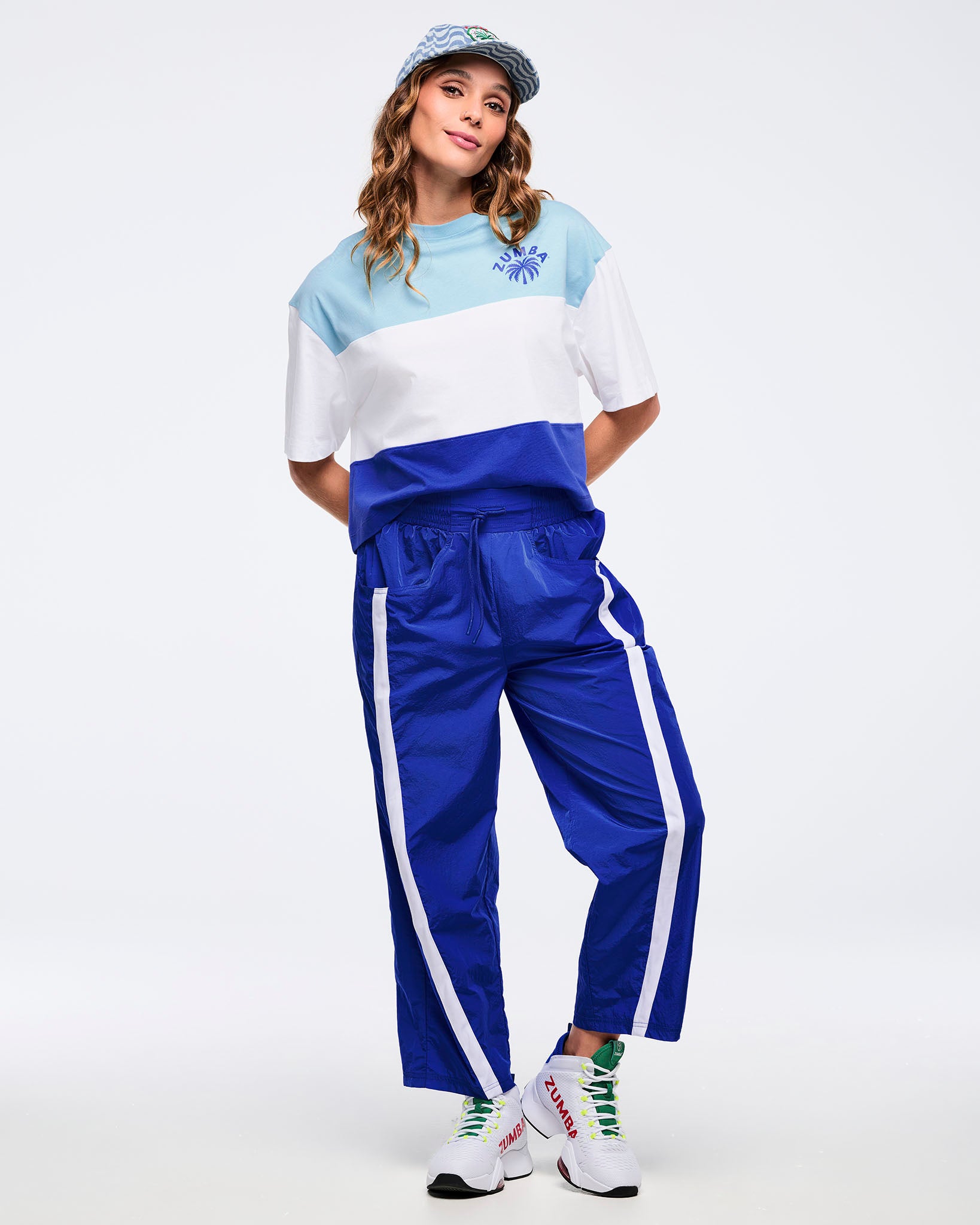 Color-block oversized T-shirt in light blue, white, and royal blue with a small Zumba palm logo, worn by a female model with bright blue track pants featuring white side stripes, a patterned baseball cap, and white high-top sneakers.