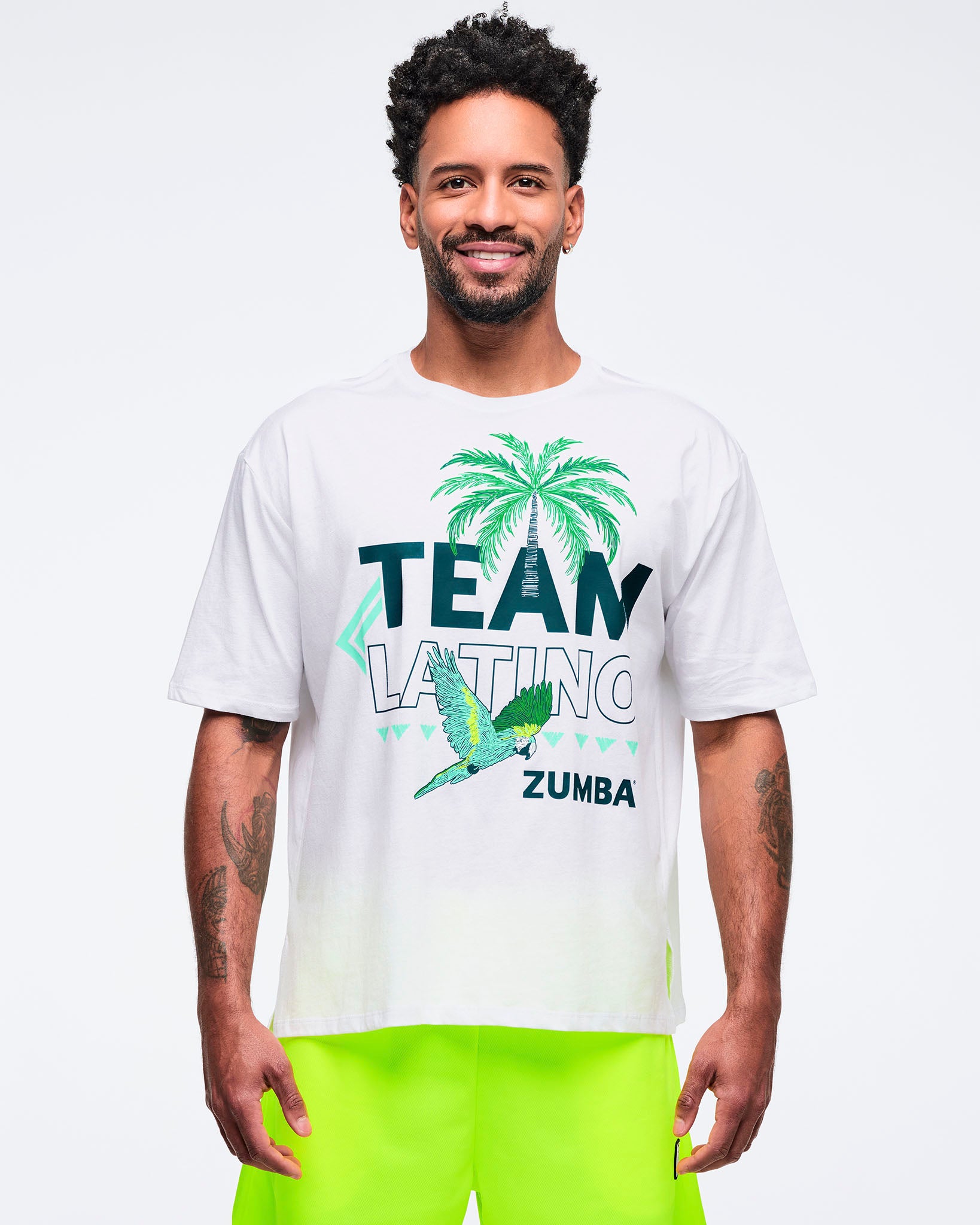 White “Team Latino” Zumba graphic T-shirt featuring a palm tree and parrot print in teal and green. Male model wears the relaxed-fit tee with neon yellow shorts on a light gray studio background.