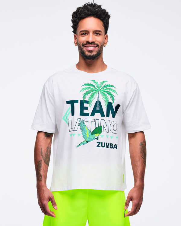 White “Team Latino” Zumba graphic T-shirt featuring a palm tree and parrot print in teal and green. Male model wears the relaxed-fit tee with neon yellow shorts on a light gray studio background.