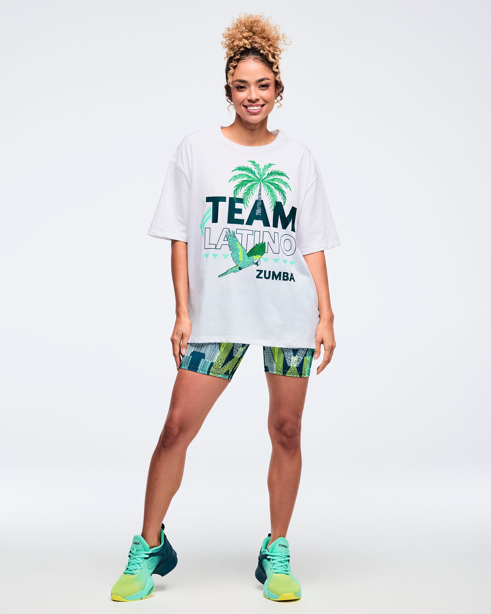 White oversized “Team Latino” Zumba graphic T-shirt with palm tree and parrot design. Female model styles it with teal-and-lime patterned biker shorts and teal gradient Zumba sneakers, standing on a light gray studio background.