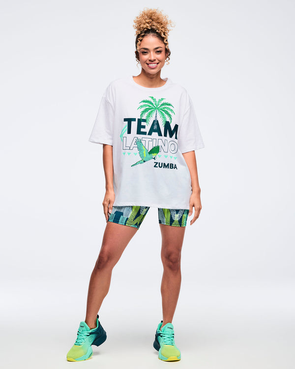 White oversized “Team Latino” Zumba graphic T-shirt with palm tree and parrot design. Female model styles it with teal-and-lime patterned biker shorts and teal gradient Zumba sneakers, standing on a light gray studio background.