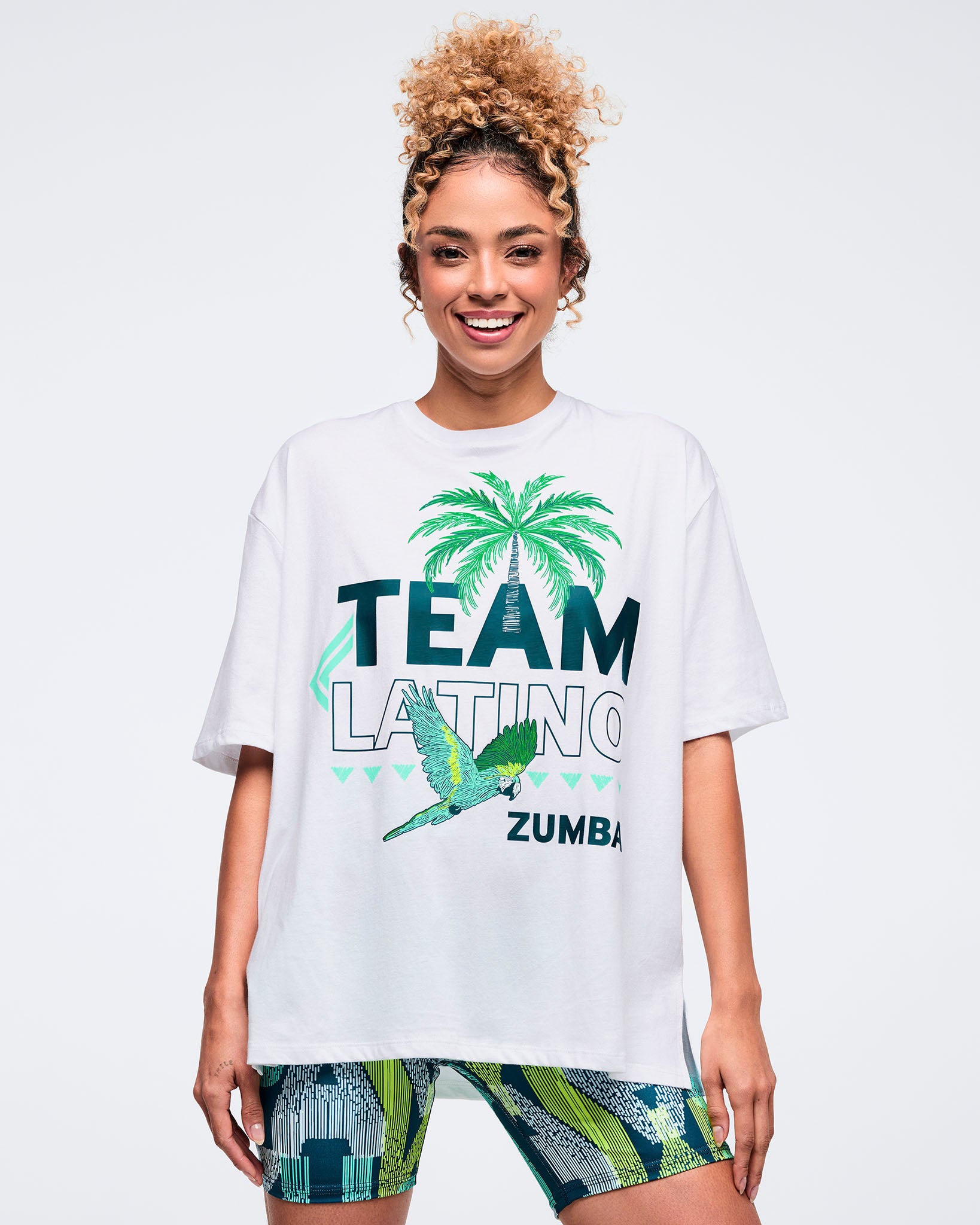 White oversized “Team Latino” Zumba graphic T-shirt with palm tree and parrot print in teal and green. Female model wears the loose, drop-shoulder tee with patterned teal-and-lime biker shorts on a light gray studio background.