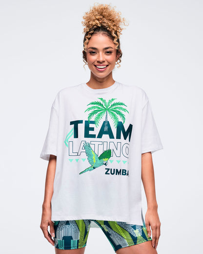 White oversized “Team Latino” Zumba graphic T-shirt with palm tree and parrot print in teal and green. Female model wears the loose, drop-shoulder tee with patterned teal-and-lime biker shorts on a light gray studio background.