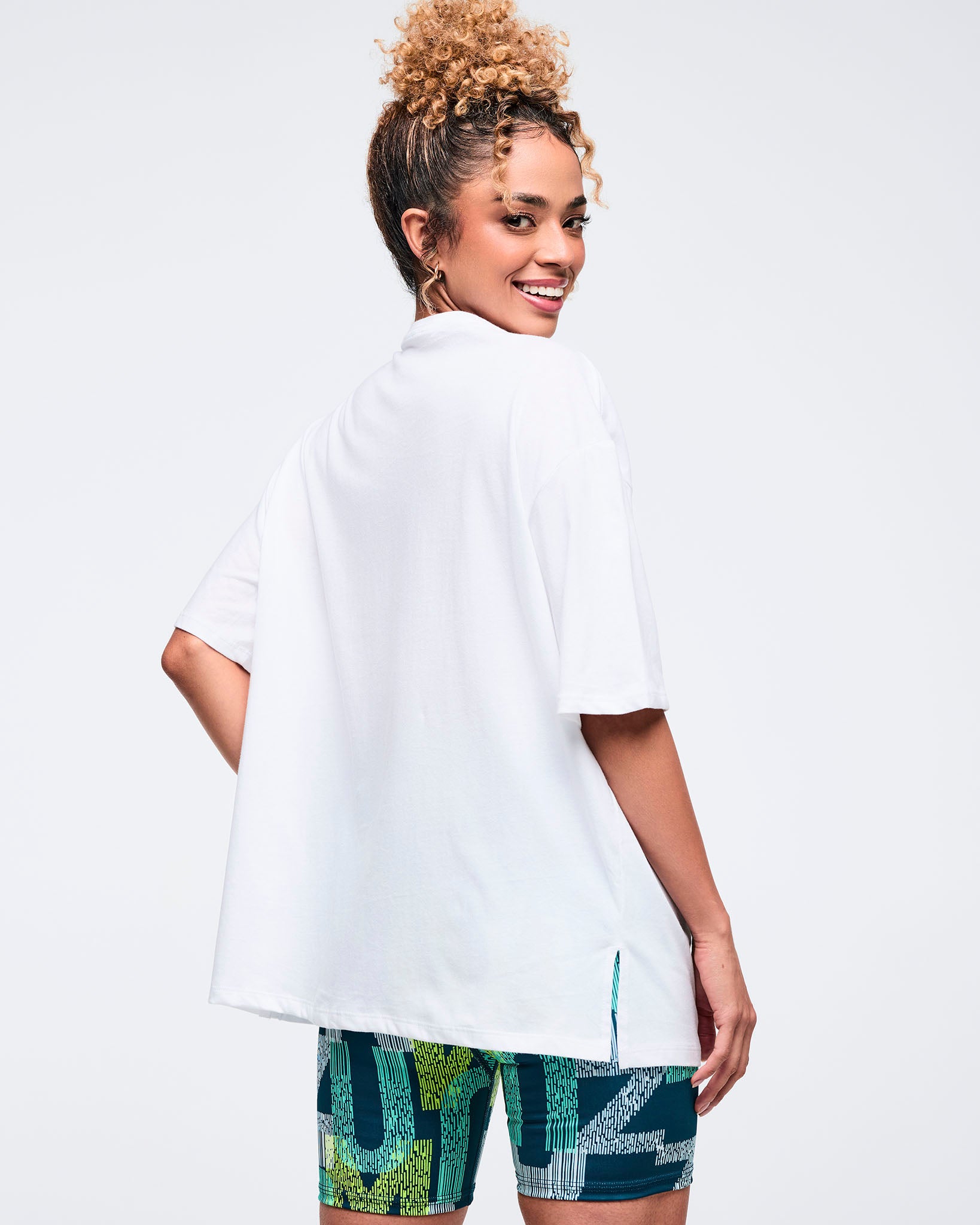 Back view of a white oversized Zumba T-shirt with a plain back, short sleeves, and long hem with side slits. Female model pairs it with teal-and-lime patterned biker shorts on a light gray studio background.