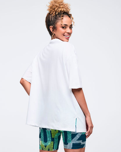 Back view of a white oversized Zumba T-shirt with a plain back, short sleeves, and long hem with side slits. Female model pairs it with teal-and-lime patterned biker shorts on a light gray studio background.